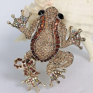 K&ouml;nnte beinhalten: Ein Frosch-Broschen-Anstecker mit goldfarbenem Metallrahmen, besetzt mit hell- und dunkelbraunen Strasssteinen. Die Augen des Frosches sind schwarz, und die F&uuml;&szlig;e haben schillernde Strasssteine. Ein dekoratives Accessoire.