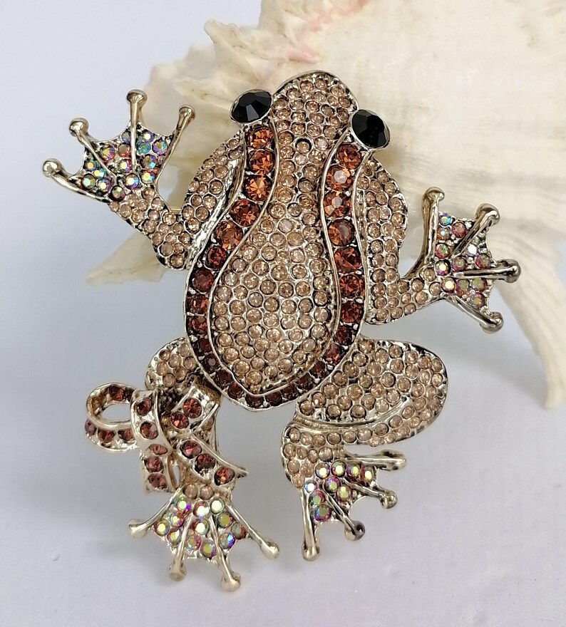 K&ouml;nnte beinhalten: Ein Frosch-Broschen-Anstecker mit goldfarbenem Metallrahmen, besetzt mit hell- und dunkelbraunen Strasssteinen. Die Augen des Frosches sind schwarz, und die F&uuml;&szlig;e haben schillernde Strasssteine. Ein dekoratives Accessoire.