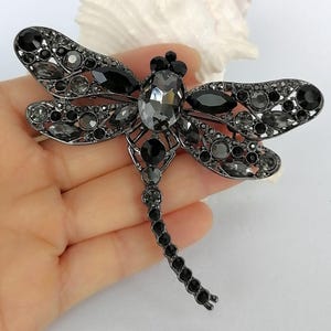 May include: A dragonfly brooch in a dark gunmetal tone, embellished with an array of black and grey crystals. The brooch has a prominent central crystal and intricate wing detailing, forming a glamorous, eye-catching accessory.