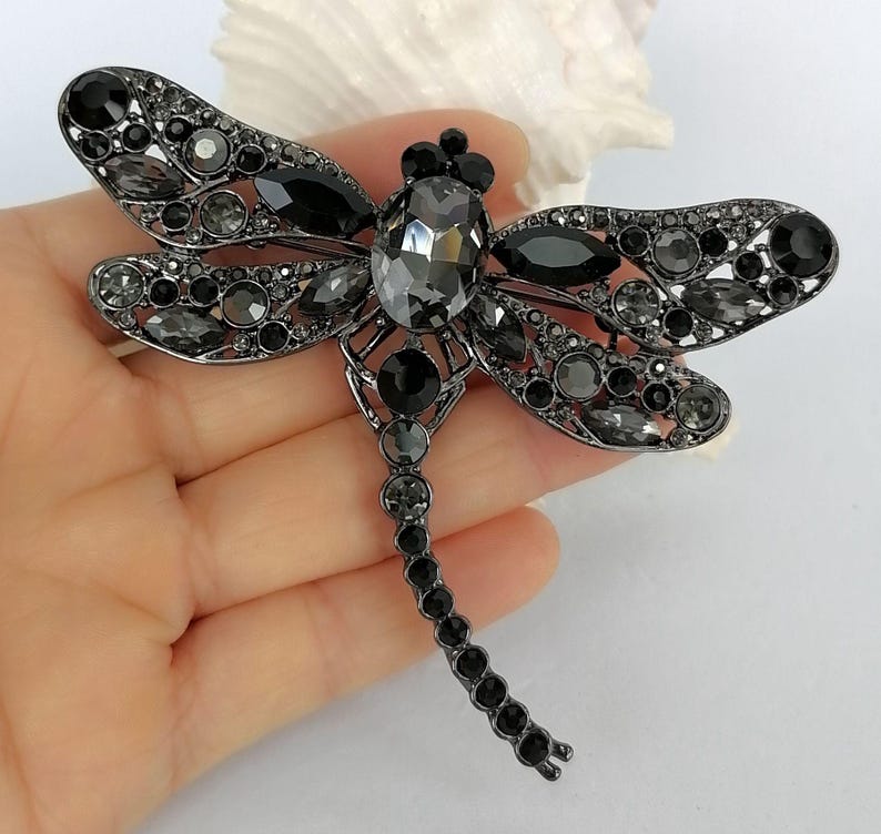 May include: A dragonfly brooch in a dark gunmetal tone, embellished with an array of black and grey crystals. The brooch has a prominent central crystal and intricate wing detailing, forming a glamorous, eye-catching accessory.