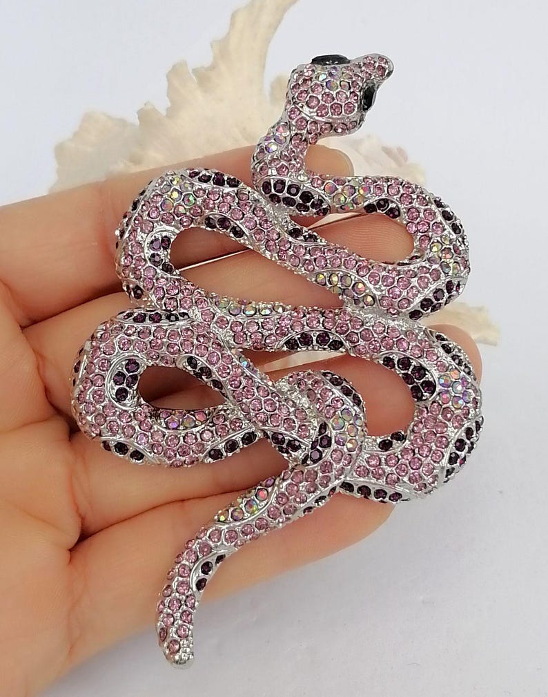 May include: A decorative snake brooch, featuring a design of coiled serpent. The brooch is adorned with pink and purple crystals, set on a silver-toned metal base. The crystals create a sparkling effect, adding to the brooch's appeal.