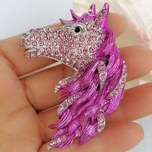 K&ouml;nnte beinhalten: Detaillierte Brosche in Form eines Pferdekopfes. Das Pferd hat eine silberfarbene Schnauze und ist mit rosa Strasssteinen besetzt. Die M&auml;hne ist leuchtend magenta, ebenfalls mit rosa Strasssteinen akzentuiert.