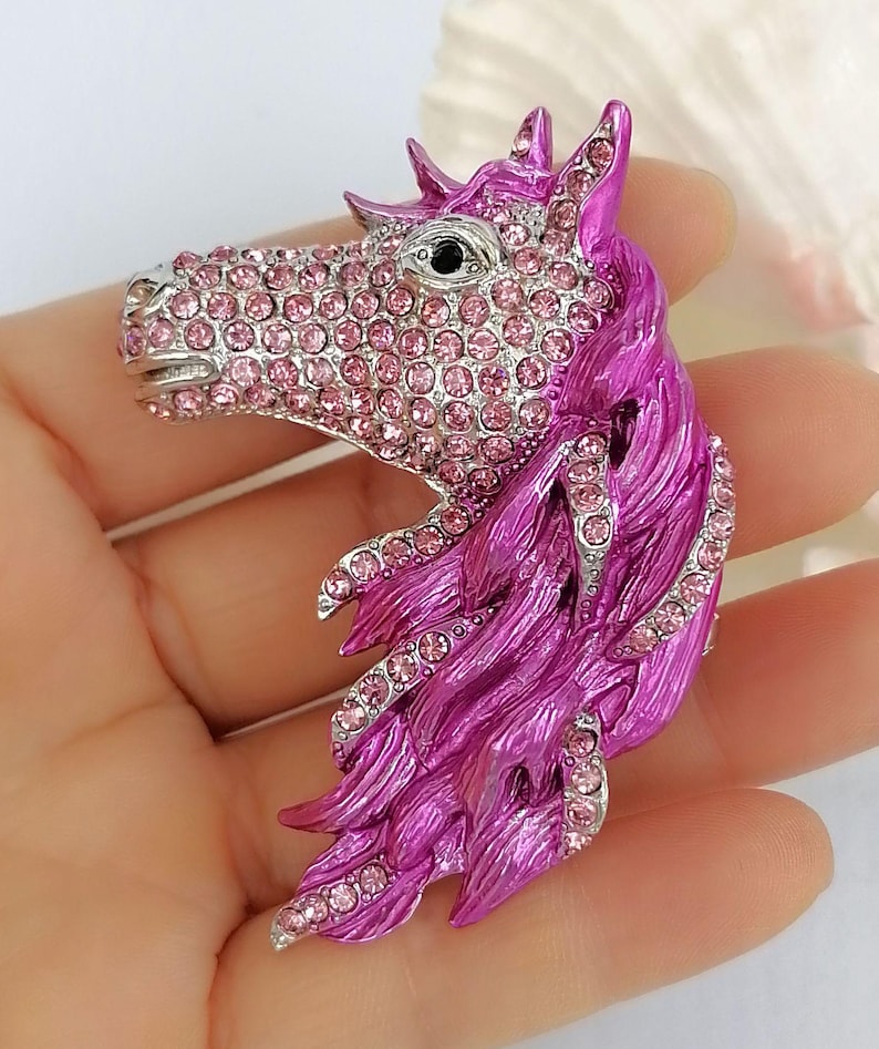K&ouml;nnte beinhalten: Detaillierte Brosche in Form eines Pferdekopfes. Das Pferd hat eine silberfarbene Schnauze und ist mit rosa Strasssteinen besetzt. Die M&auml;hne ist leuchtend magenta, ebenfalls mit rosa Strasssteinen akzentuiert.