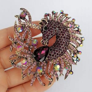 May include: A detailed brooch featuring a purple unicorn design. The unicorn is encrusted with small, dark purple rhinestones, with a large, teardrop-shaped, dark purple gem. The brooch has gold-colored accents and iridescent crystals.