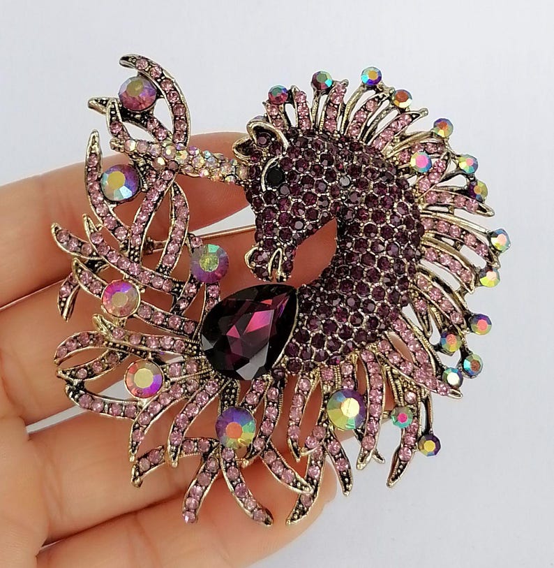 May include: A detailed brooch featuring a purple unicorn design. The unicorn is encrusted with small, dark purple rhinestones, with a large, teardrop-shaped, dark purple gem. The brooch has gold-colored accents and iridescent crystals.