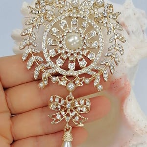 May include: A gold-tone brooch featuring a circular frame adorned with clear rhinestones and small white faux pearls. The design includes a central pearl, a wreath-like border, a dangling bow accent, and a single teardrop-shaped faux pearl pendant.
