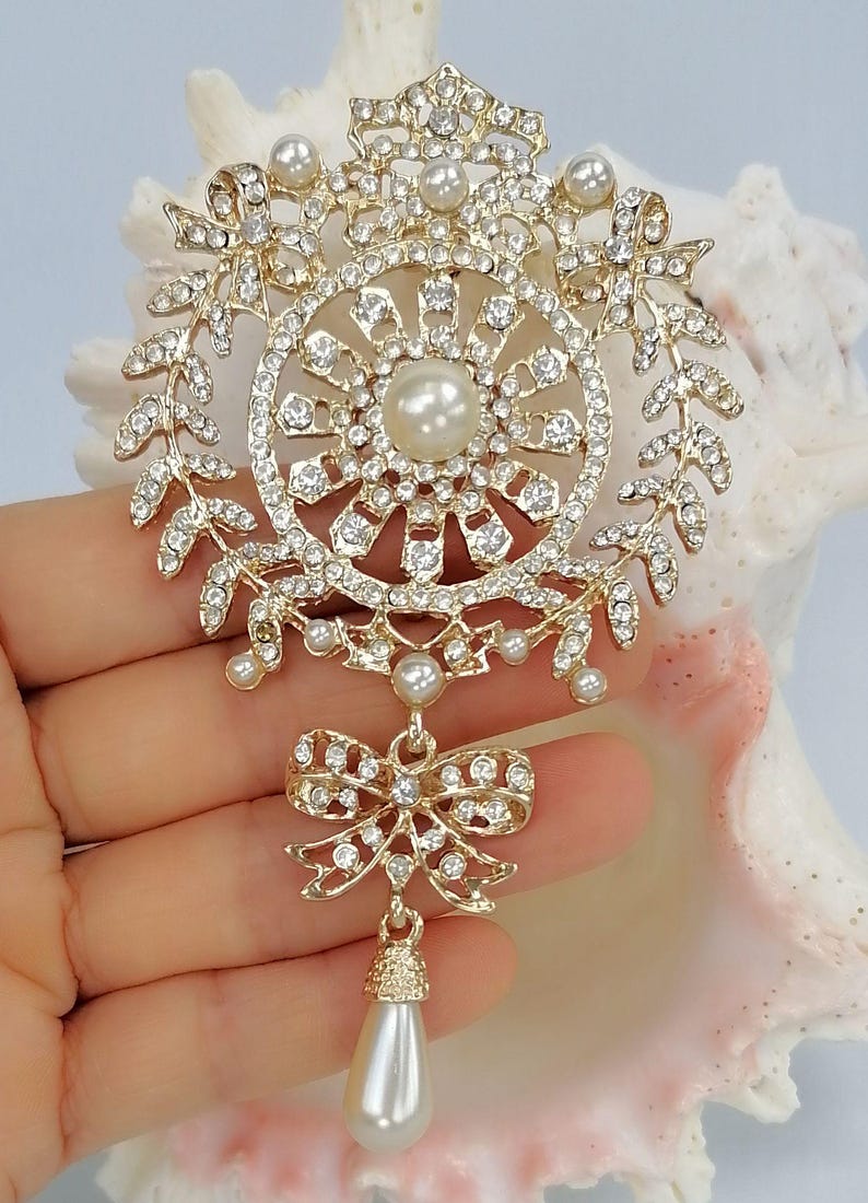 May include: A gold-tone brooch featuring a circular frame adorned with clear rhinestones and small white faux pearls. The design includes a central pearl, a wreath-like border, a dangling bow accent, and a single teardrop-shaped faux pearl pendant.