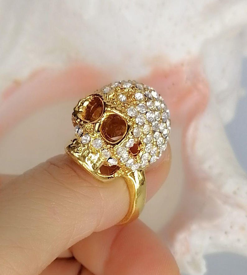 Op de afbeelding: Een goudkleurige ring met een driedimensionaal schedelontwerp. De schedel is ingelegd met talloze kleine, heldere, sprankelende steentjes die de schedel en jukbeenderen bedekken, terwijl de oogkassen en de neusholte glad en hol blijven.