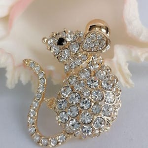 May include: A gold-toned mouse brooch encrusted with clear crystals. The mouse has a black eye and a curved tail. The brooch is set against a soft, blurred background, highlighting the sparkling details of the jewelry.