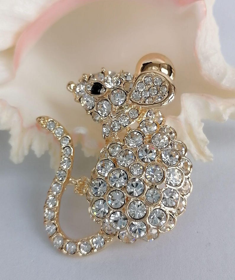 May include: A gold-toned mouse brooch encrusted with clear crystals. The mouse has a black eye and a curved tail. The brooch is set against a soft, blurred background, highlighting the sparkling details of the jewelry.