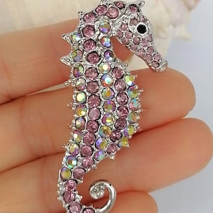 May include: A silver-toned seahorse brooch encrusted with pink and iridescent crystals. The seahorse has a detailed design with a curled tail and a small black eye. The brooch is a decorative accessory.