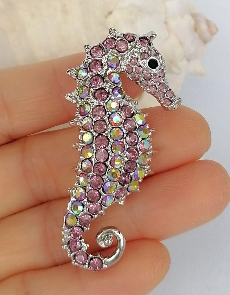 May include: A silver-toned seahorse brooch encrusted with pink and iridescent crystals. The seahorse has a detailed design with a curled tail and a small black eye. The brooch is a decorative accessory.
