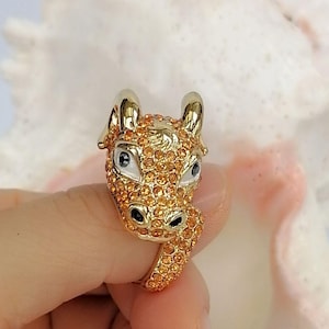 May include: A gold-toned ring featuring a sculpted animal head design, likely a bull or ox, covered in small orange crystals. The animal has smooth gold horns, white enamel around the eyes with black pupils, and black nostrils, with the band also encrusted in orange crystals.