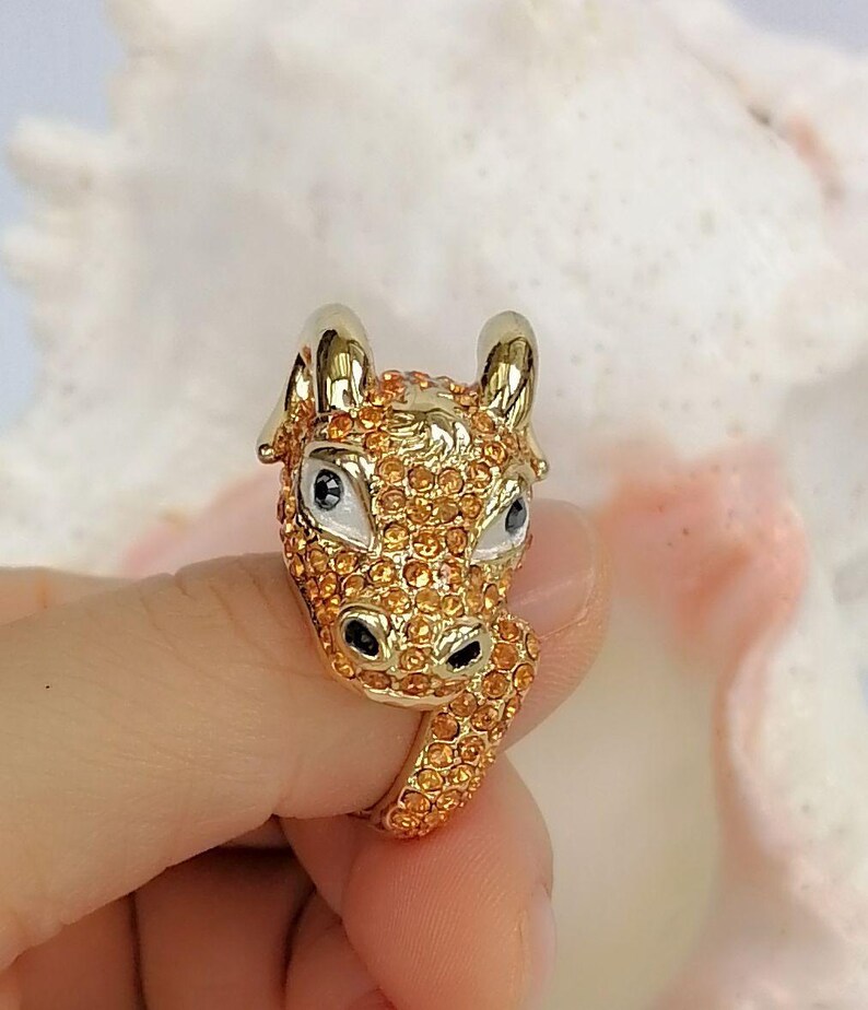 May include: A gold-toned ring featuring a sculpted animal head design, likely a bull or ox, covered in small orange crystals. The animal has smooth gold horns, white enamel around the eyes with black pupils, and black nostrils, with the band also encrusted in orange crystals.