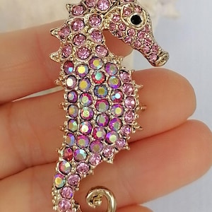 May include: A gold-toned seahorse brooch encrusted with pink and iridescent crystals. The seahorse has a curled tail and a detailed head with a black eye. The brooch is a decorative accessory.