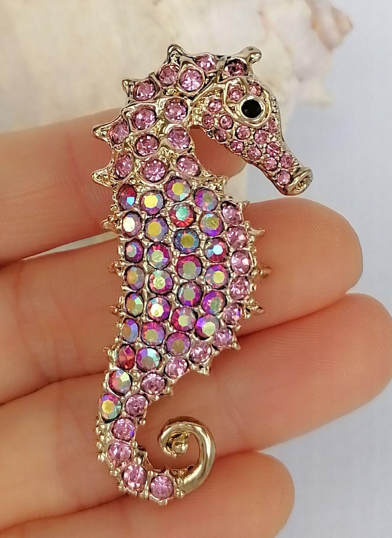 May include: A gold-toned seahorse brooch encrusted with pink and iridescent crystals. The seahorse has a curled tail and a detailed head with a black eye. The brooch is a decorative accessory.