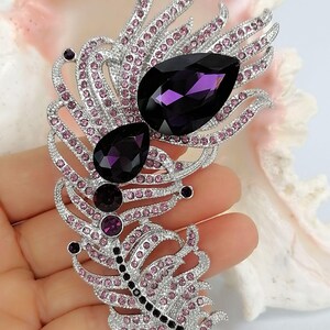 May include: A silver-toned feather brooch featuring two large, dark purple, pear-shaped crystals, two smaller round purple crystals, and numerous tiny light pink rhinestones embedded throughout the intricate metal frame of the feather design.