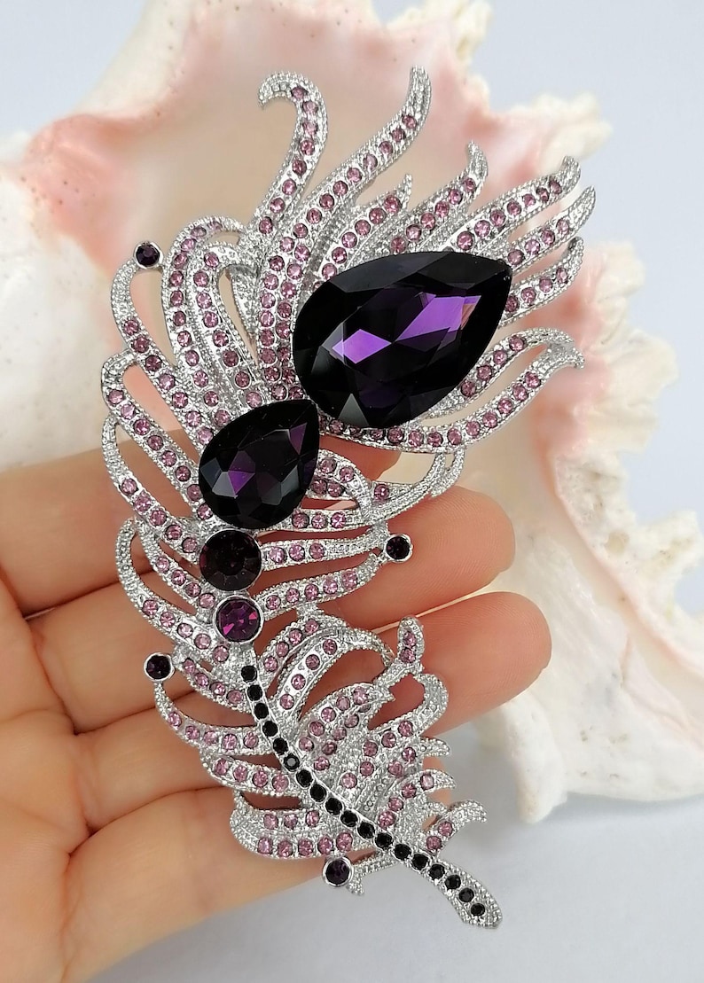 May include: A silver-toned feather brooch featuring two large, dark purple, pear-shaped crystals, two smaller round purple crystals, and numerous tiny light pink rhinestones embedded throughout the intricate metal frame of the feather design.