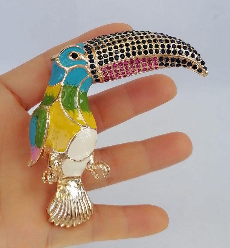 May include: A colorful toucan brooch with a gold-tone metal base. The bird features a black and pink jeweled beak, and a body painted in shades of blue, green, yellow, and white. The brooch is a decorative accessory.
