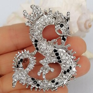 Puede incluir: Broche de drag&oacute;n plateado, incrustado con pedrer&iacute;a transparente y negra. El drag&oacute;n est&aacute; curvado en una pose estilizada, con escamas y rasgos detallados. Un accesorio decorativo.