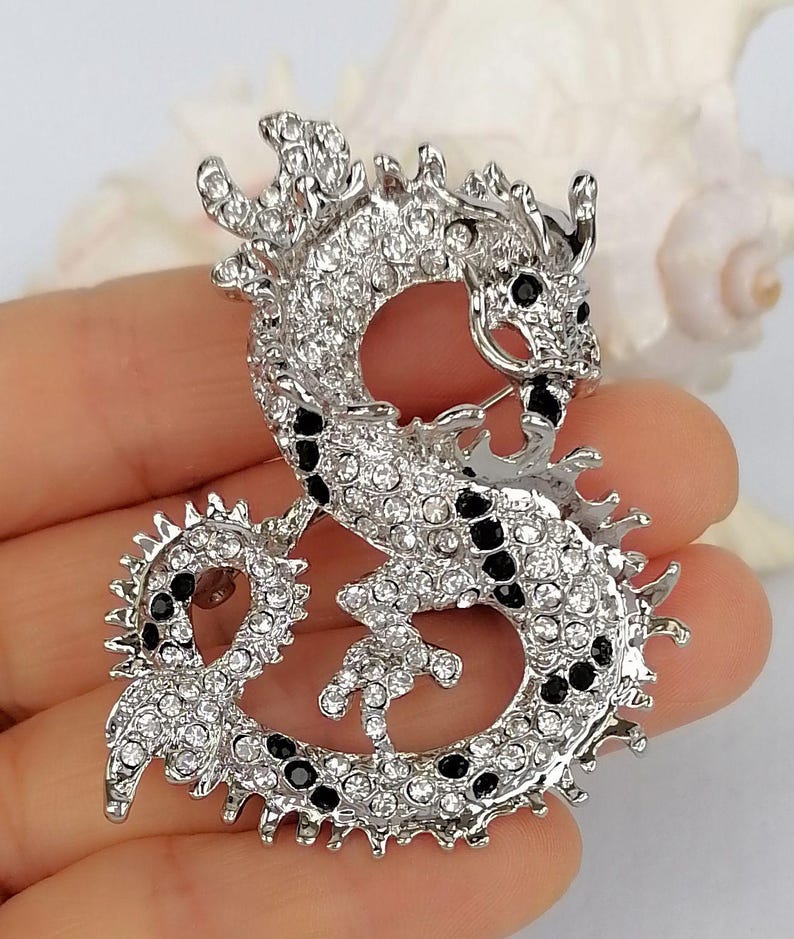 Puede incluir: Broche de drag&oacute;n plateado, incrustado con pedrer&iacute;a transparente y negra. El drag&oacute;n est&aacute; curvado en una pose estilizada, con escamas y rasgos detallados. Un accesorio decorativo.