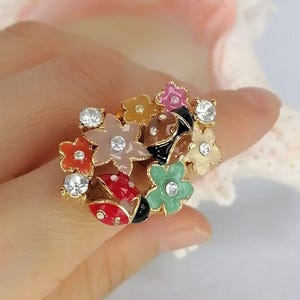 May include: A gold-tone ring featuring a cluster of colorful enamel flowers in shades of pink, orange, beige, and green, accented with clear rhinestones and two ladybug charms with red and brown enamel details and black spots.