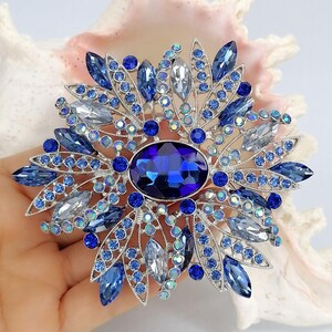 Op de afbeelding: Een zilverkleurige broche met een grote, centrale, ovaalvormige diepblauwe kristal, omringd door een radiale opstelling van kleinere marquise-geslepen lichtblauwe, donkerblauwe en heldere kristallen, geaccentueerd met kleine ronde blauwe strasssteentjes.