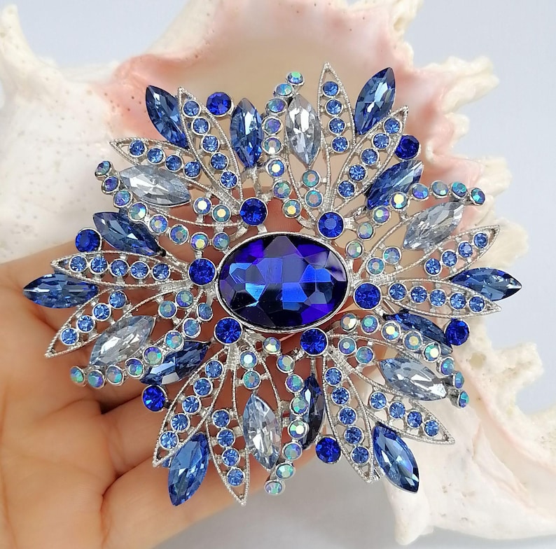 Op de afbeelding: Een zilverkleurige broche met een grote, centrale, ovaalvormige diepblauwe kristal, omringd door een radiale opstelling van kleinere marquise-geslepen lichtblauwe, donkerblauwe en heldere kristallen, geaccentueerd met kleine ronde blauwe strasssteentjes.