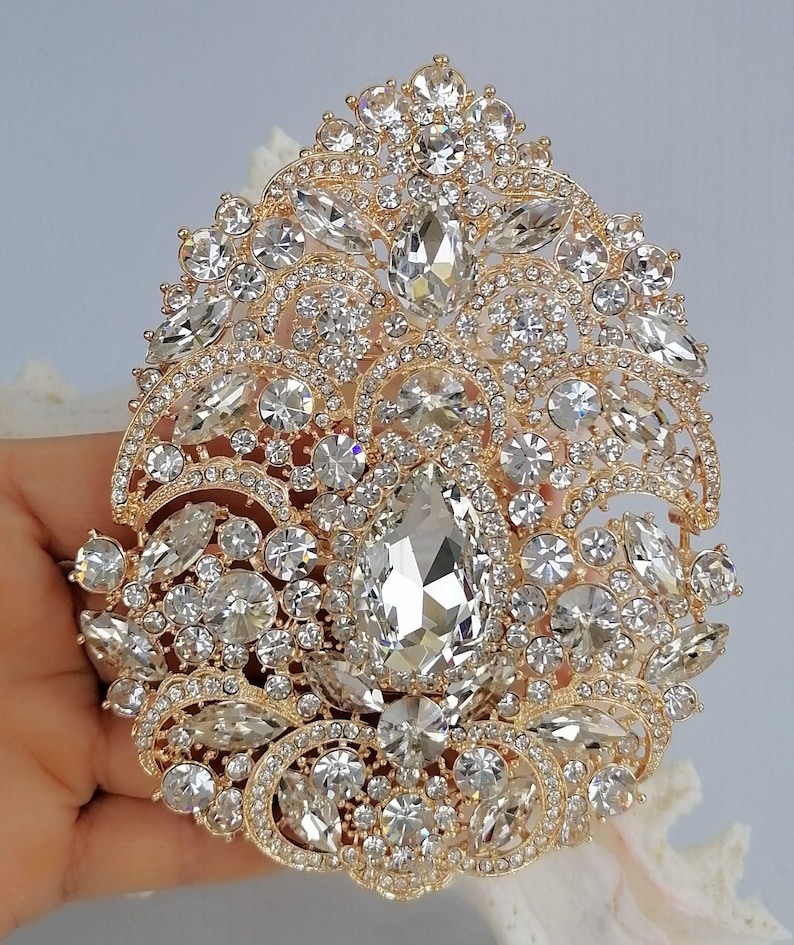 May include: A gold-toned, ornate brooch with a large, pear-shaped central stone and numerous smaller, clear crystals. The brooch has a detailed, filigree design and is likely intended for formal wear or special occasions.