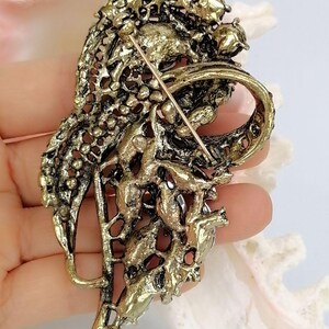 May include: An antique gold-toned metal brooch featuring an intricate, openwork filigree design with a textured, weathered finish. The back of the piece displays a secure pin mechanism with a long, thin metal needle extending across the center.