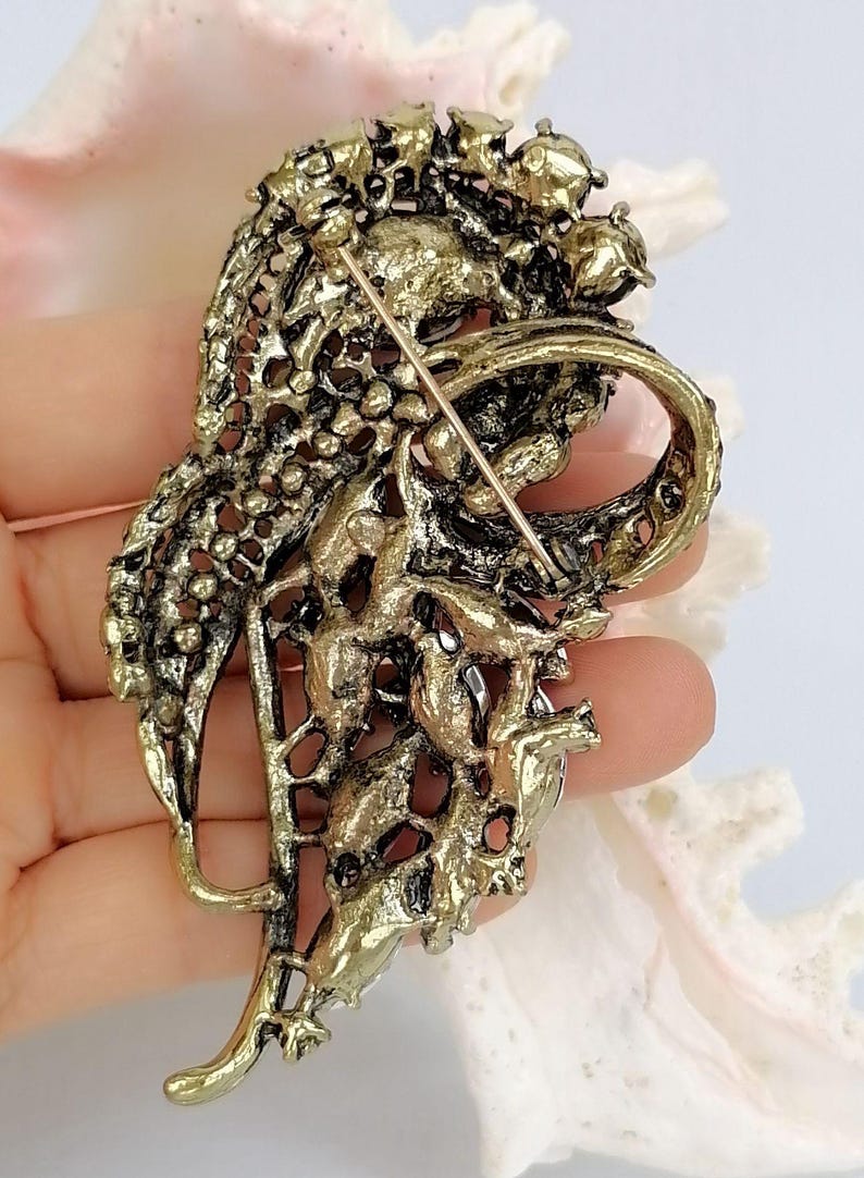 May include: An antique gold-toned metal brooch featuring an intricate, openwork filigree design with a textured, weathered finish. The back of the piece displays a secure pin mechanism with a long, thin metal needle extending across the center.