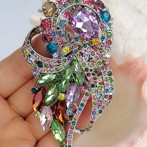 May include: A silver-tone brooch featuring a swirling, organic design encrusted with multi-colored faceted crystals. The piece includes large marquise and pear-shaped stones in shades of green, purple, and red, accented by smaller round crystals.