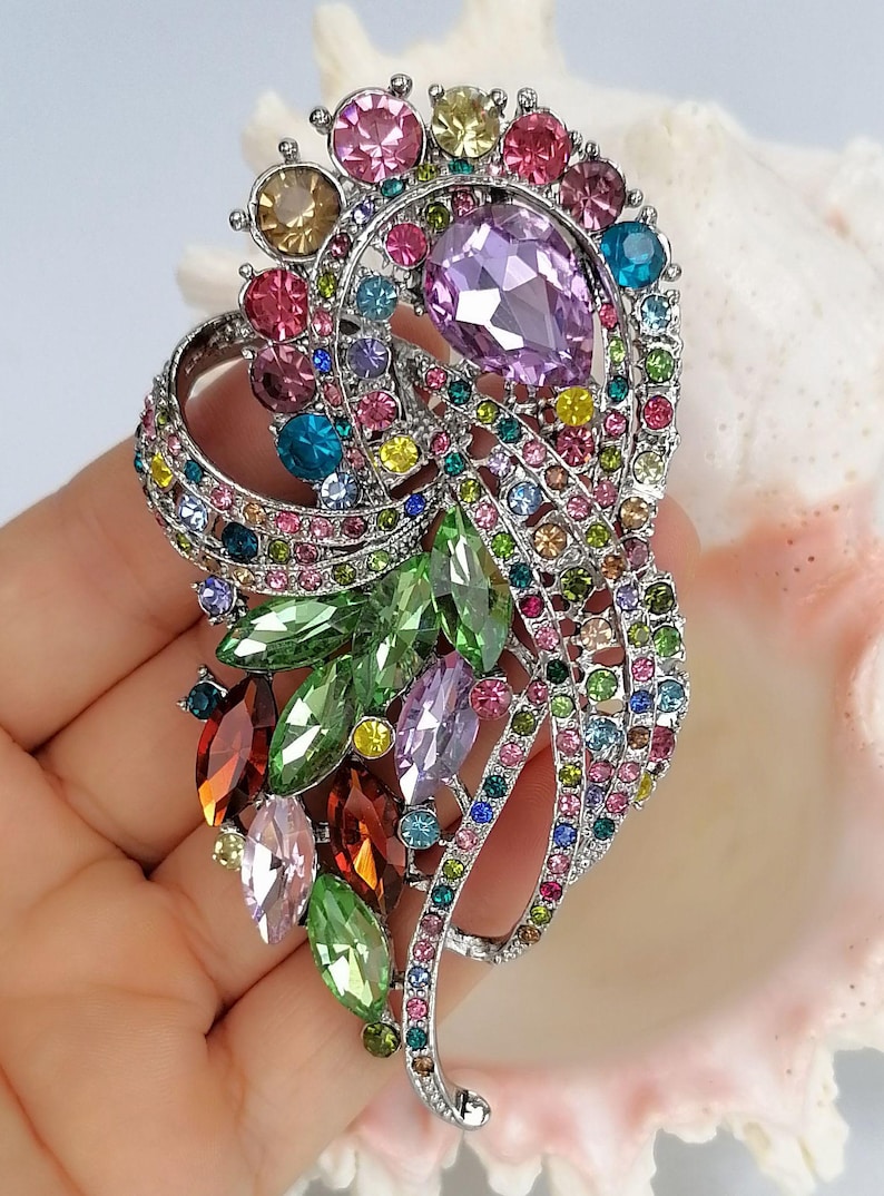 May include: A silver-tone brooch featuring a swirling, organic design encrusted with multi-colored faceted crystals. The piece includes large marquise and pear-shaped stones in shades of green, purple, and red, accented by smaller round crystals.