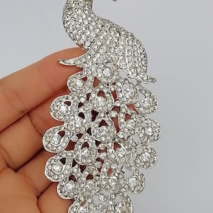 May include: A silver-toned peacock brooch, encrusted with numerous clear rhinestones. The brooch features a detailed design of a peacock with its tail feathers spread. The brooch is held in a hand against a plain background.