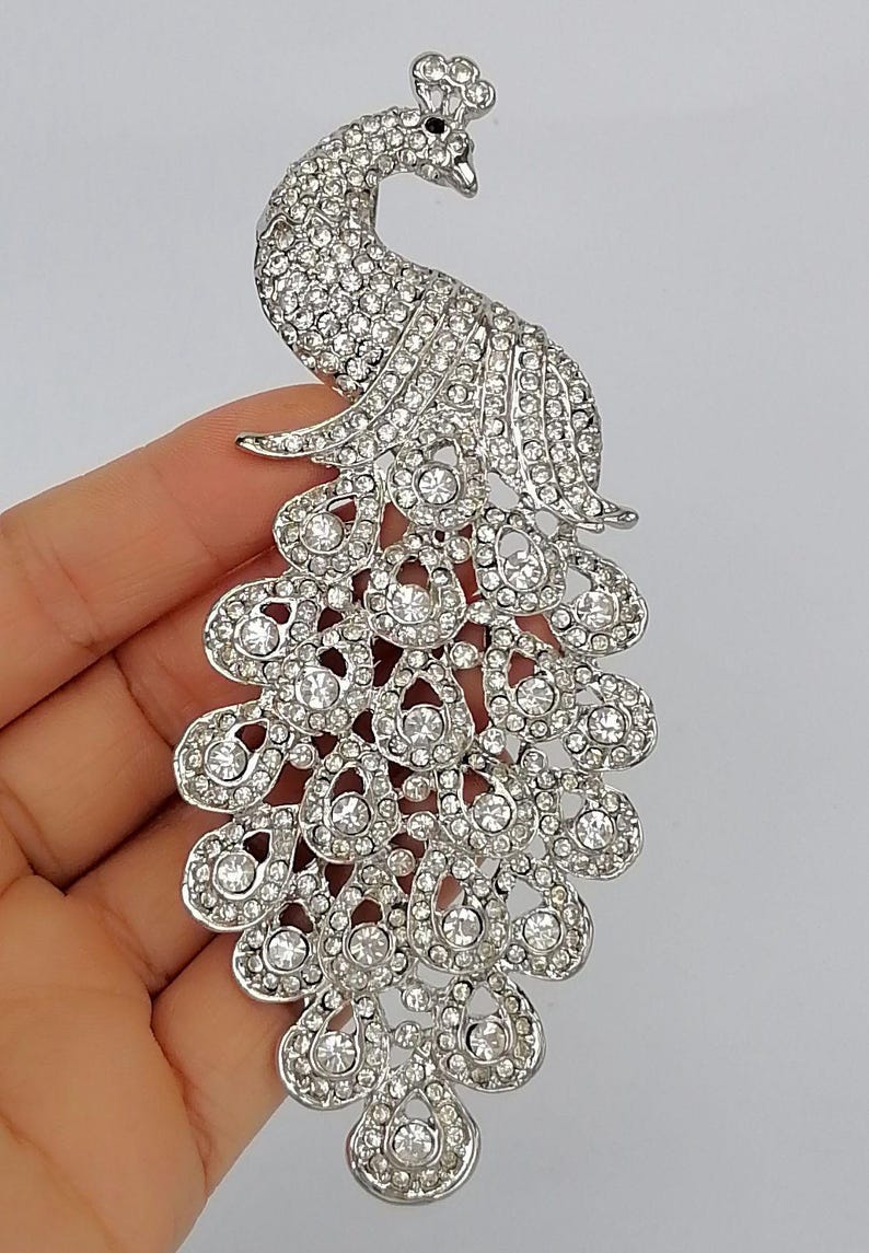 May include: A silver-toned peacock brooch, encrusted with numerous clear rhinestones. The brooch features a detailed design of a peacock with its tail feathers spread. The brooch is held in a hand against a plain background.