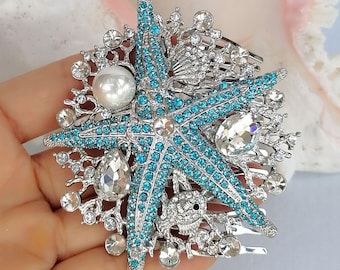 May include: A silver-toned hair comb featuring a central starfish motif encrusted with bright blue crystals. The design includes clear crystal accents, a single white faux pearl, and a small silver crab figure, all set against a metallic base.