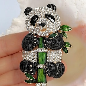 May include: A panda brooch featuring a head and body covered in clear sparkling rhinestones, black textured enamel limbs, and ears. The panda is holding a green enameled bamboo stalk with small green leaves and clear rhinestone accents.