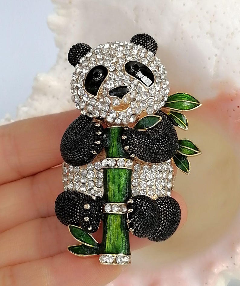 May include: A panda brooch featuring a head and body covered in clear sparkling rhinestones, black textured enamel limbs, and ears. The panda is holding a green enameled bamboo stalk with small green leaves and clear rhinestone accents.