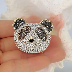 May include: A panda-shaped brooch featuring a gold-tone metal frame encrusted with clear white and black crystals. The design depicts the panda's face with black crystal patches around the eyes and ears, and a small black bead for the nose.