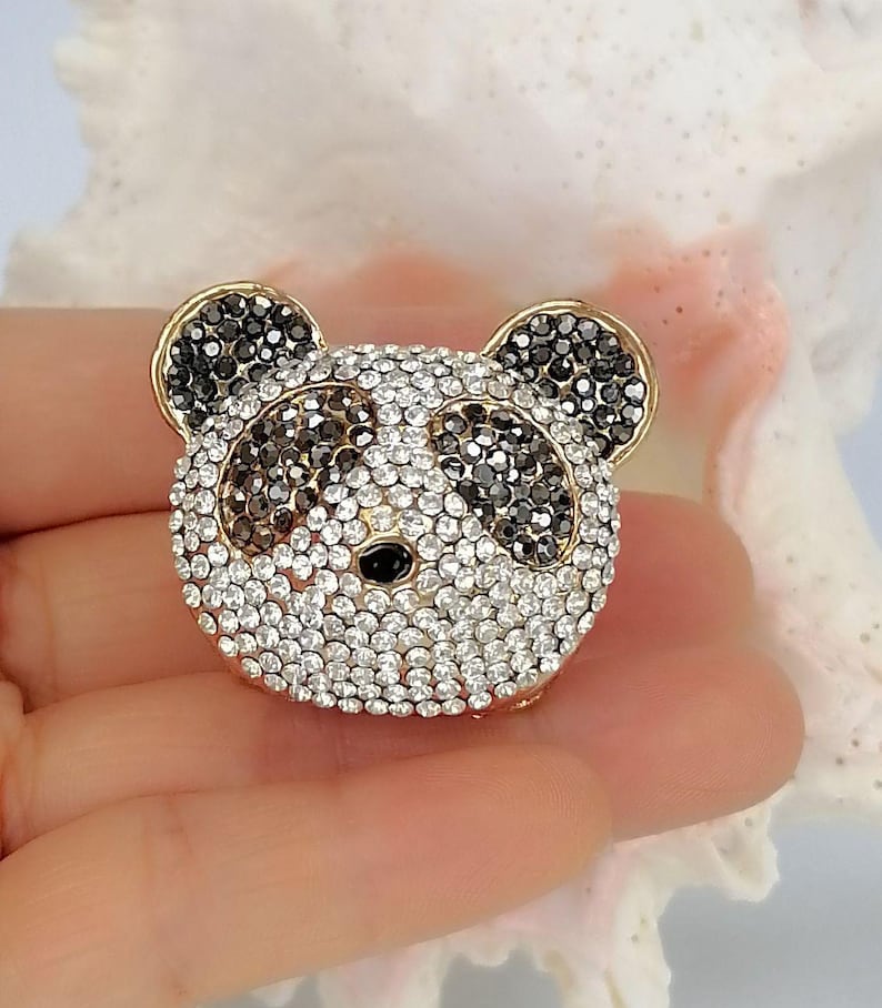 May include: A panda-shaped brooch featuring a gold-tone metal frame encrusted with clear white and black crystals. The design depicts the panda's face with black crystal patches around the eyes and ears, and a small black bead for the nose.