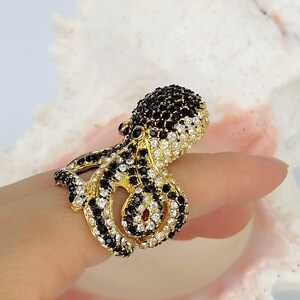 Peut inclure: Une bague dor&eacute;e en forme de pieuvre, pr&eacute;sentant un design d&eacute;taill&eacute; incrust&eacute; de nombreuses petites pierres &eacute;tincelantes noires et blanches transparentes qui recouvrent toute la surface de la t&ecirc;te et des tentacules enroul&eacute;s de la cr&eacute;ature.