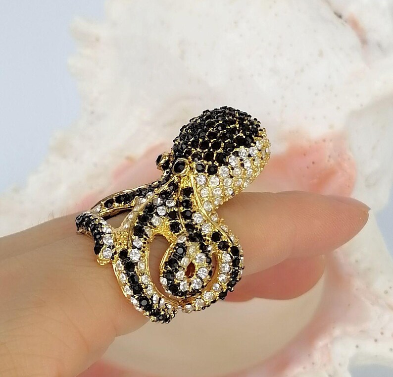 Peut inclure: Une bague dor&eacute;e en forme de pieuvre, pr&eacute;sentant un design d&eacute;taill&eacute; incrust&eacute; de nombreuses petites pierres &eacute;tincelantes noires et blanches transparentes qui recouvrent toute la surface de la t&ecirc;te et des tentacules enroul&eacute;s de la cr&eacute;ature.