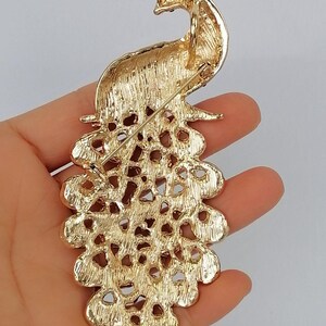 May include: Gold-toned peacock brooch with intricate detailing. The brooch features a detailed peacock design with a crown and a fanned tail. The brooch is held in a hand, showcasing the back of the pin.