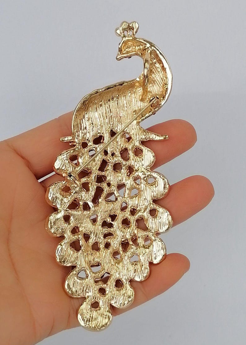 May include: Gold-toned peacock brooch with intricate detailing. The brooch features a detailed peacock design with a crown and a fanned tail. The brooch is held in a hand, showcasing the back of the pin.