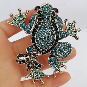K&ouml;nnte beinhalten: Eine goldfarbene Froschbrosche, besetzt mit teal- und gr&uuml;nen Strasssteinen. Der Frosch hat schwarze Augen und ausgestreckte Gliedma&szlig;en. Die Brosche ist ein skurriles Accessoire, perfekt, um jedem Outfit einen Hauch von von der Natur inspiriertem Glanz zu verleihen.
