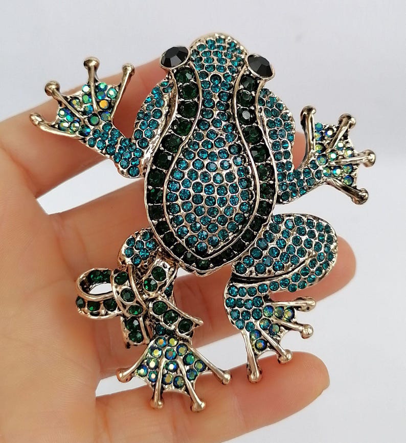 K&ouml;nnte beinhalten: Eine goldfarbene Froschbrosche, besetzt mit teal- und gr&uuml;nen Strasssteinen. Der Frosch hat schwarze Augen und ausgestreckte Gliedma&szlig;en. Die Brosche ist ein skurriles Accessoire, perfekt, um jedem Outfit einen Hauch von von der Natur inspiriertem Glanz zu verleihen.