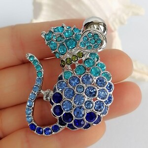May include: A silver-toned mouse brooch encrusted with blue and teal crystals. The mouse has a rounded body and a long, curved tail. The brooch features a silver ball for the head and is set against a soft background.