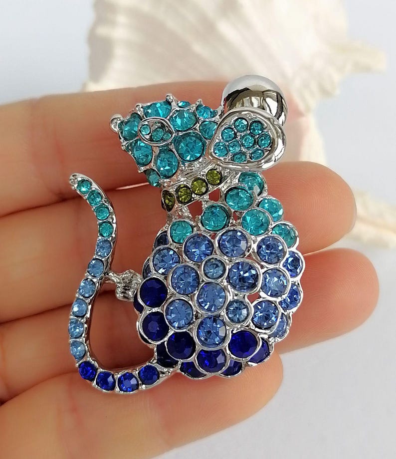 May include: A silver-toned mouse brooch encrusted with blue and teal crystals. The mouse has a rounded body and a long, curved tail. The brooch features a silver ball for the head and is set against a soft background.