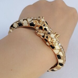 May include: A gold-toned hinged bangle bracelet featuring two giraffe heads meeting at the center. The giraffe pattern is rendered in black and cream enamel, with small clear crystals embedded along the side of the band near the giraffe heads.