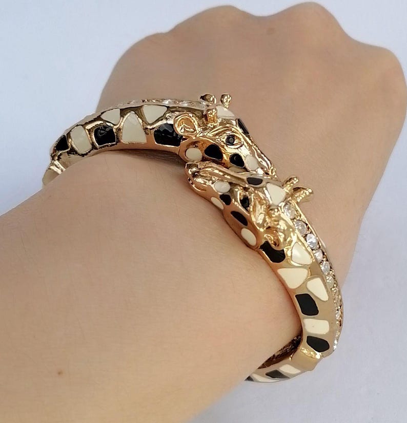 May include: A gold-toned hinged bangle bracelet featuring two giraffe heads meeting at the center. The giraffe pattern is rendered in black and cream enamel, with small clear crystals embedded along the side of the band near the giraffe heads.